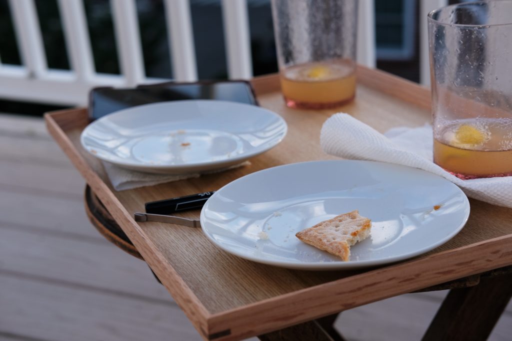 image of a tray on a table on a wooden deck. There is one empty plate next to a cell phone, and another plate with a half eaten pastry next to a ben, and two nearly empty water glasses with lemon