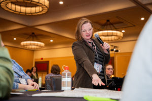 A person standing at a table holding a microphone and pointing at their large sticky note on the table.